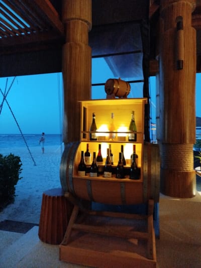 A barrel-shaped wine rack featuring several bottles of wine and a small wooden barrel is displayed prominently in the foreground, situated on a beach in Kendhoo, Maldives. The wine rack is illuminated, highlighting the bottles of various shapes and colors, some with labels. In the background, the ocean meets the sandy beach, with a person standing near the water's edge. The scene is set during dusk or twilight, with the sky displaying a deep blue hue. The overall atmosphere suggests a tranquil and inviting setting, likely at a resort or a beachfront establishment.