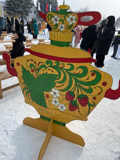 The image displays an outdoor winter scene, captured sideways such that the ground appears on the right and left sides, and vertical elements are horizontal. If rotated 90 degrees counter-clockwise to its intended upright orientation, the scene shows a large, brightly painted wooden cutout resembling a teapot or samovar, standing prominently in a snow-covered landscape.
The central object is a large, flat, freestanding wooden cutout, predominantly golden yellow, with intricate folk art-style decorations. It depicts a traditional Russian teapot or samovar. The body of the object is decorated with flowing green vines and large green leaves, possibly strawberry leaves. Scattered among the foliage are clusters of red strawberries with small green caps, white flowers with yellow centers, and small red flowers. The design also features numerous small white and red dots, adding to the decorative detail. Red borders outline parts of the teapot, including its handle (on the left, if upright) and spout (on the far left, if upright). The cutout appears to be supported by wooden braces or stands, visible at the bottom right of the image (or bottom, if upright).
Numerous people are visible in the midground and background, all dressed in heavy winter clothing such as dark puffer coats, hats, and scarves, indicating cold weather. Some individuals are seen standing near the large cutout, while others are further back, forming loose groups or lines. One person in the foreground, wearing a black coat and black hat, is visible near the base of the cutout. In the background, slightly to the left, another person holds up a circular yellow object with radiating lines, resembling a stylized sun, possibly a prop for an event.
The ground is uniformly covered in a thick layer of white snow. In the background, there are bare deciduous trees and some evergreen trees. Small flags or banners, predominantly red, white, and blue, are strung up in the left background, suggesting a festive or public gathering. The sky is bright and overcast, characteristic of a winter day.
The overall impression is that of an outdoor winter festival or celebration, likely with a focus on Russian culture, given the traditional folk art style of the decorative teapot and the presence of what appears to be a sun effigy (often associated with Maslenitsa celebrations). No visible text is present in the image.