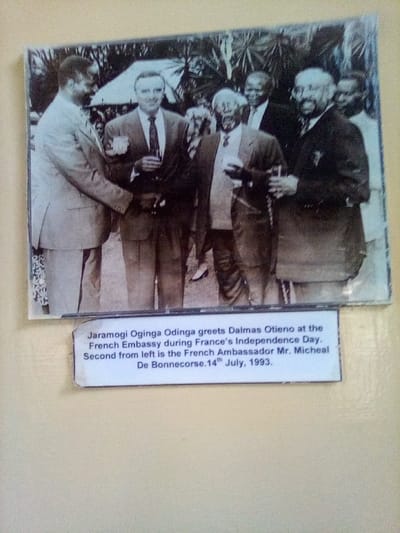 A photograph of a black and white image is displayed on a light-colored wall in South Sakwa ward, Kenya. The black and white image depicts an outdoor gathering of at least five adult men, formally dressed in suits and ties, on July 14, 1993, at the French Embassy during France's Independence Day. In the foreground, Jaramogi Oginga Odinga, wearing a light-colored suit, extends his right hand to shake hands with Dalmas Otieno, dressed in a darker suit, who is holding a glass in his left hand. To the right of Odinga, slightly behind him, stands the French Ambassador Mr. Micheal De Bonnecorse, also in a dark suit and holding a glass. Additional men, similarly attired and some holding glasses, are visible in the background. The environment features dense foliage and a partial light-colored canopy or tent structure.