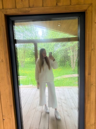 A young woman is prominently featured in the reflection of a large window or glass door, appearing to be taking a selfie. The image is captured from inside a wooden structure, looking outwards.

The woman, with long, light brown hair, is dressed in a casual, monochromatic outfit, consisting of a light-colored (likely white or cream) long-sleeved top and matching wide-leg trousers, paired with white sneakers. She holds a smartphone, specifically an iPhone with a distinctive case, in her right hand, pointing it towards herself as if capturing her reflection. A dark bracelet is visible on her left wrist. Her expression is neutral, focused on the phone screen.

The immediate interior surrounding the window is constructed from warm-toned wooden panels, forming both the wall and the window frame. The window frame itself is dark, likely black. The woman's reflection shows her standing on a light-colored wooden deck or floor.

Beyond the glass, a vibrant outdoor scene unfolds. Lush green trees and a grassy field adorned with small yellow flowers are clearly visible, indicating a natural, possibly rural, setting during spring or summer. A light-colored wooden path or deck is also discernible outside the window, extending into the verdant landscape.

The time of day is daytime, characterized by bright, natural light. The weather appears clear. The overall setting suggests a peaceful, natural environment, possibly a cabin or country house in Vozhoiskii, Russia. No visible text is present in the image.