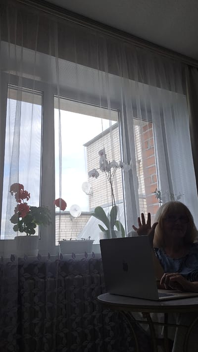 The image shows a woman sitting at a table in front of a laptop with her hand raised in a wave. She is wearing glasses and a blue top. The scene appears to be indoors, with a window behind her that looks out onto a brick building and a cloudy sky. There are plants on the windowsill, including an orchid. The setting is a room with sheer curtains and a radiator cover with a floral pattern. The woman is likely in her home, possibly in Lubnya, Russia, as per the prompt.