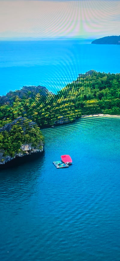 An aerial view captures a serene body of turquoise water surrounding a lush, green island with rocky outcrops. In the foreground, a small, flat raft with a bright red canopy floats on the water. The raft appears to be carrying some unidentified objects. The island is densely vegetated with trees and foliage, and a small sandy beach is visible on its shore. In the distance, the vast expanse of the ocean stretches to meet the horizon, with faint outlines of more distant landmasses. The water is calm, reflecting the clear blue sky. The location is not Lippstadt, Germany.