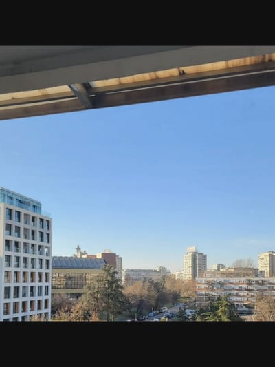 The image is an outdoor, daytime shot taken from an elevated position, likely a balcony or window, looking out over a cityscape. The foreground features the underside of a structure, possibly an awning or roof overhang, with visible wooden slats and metal supports.
The main scene depicts a clear, bright blue sky with a faint contrail from an aircraft. Below the sky, a modern white building with numerous glass-paneled windows and balconies dominates the left side of the frame. To the right of this building, a longer structure with a reflective, glass-covered facade and a sloped roof is visible. Further into the background, trees with bare branches are interspersed with apartment buildings of varying architectural styles and heights. Some buildings are tall and slender, while others are wider and appear to be older blocks of flats with numerous small windows. A street with parked cars is visible between the trees and buildings. The overall impression is an urban environment during the day, likely in a temperate climate during the late autumn or winter due to the bare trees.