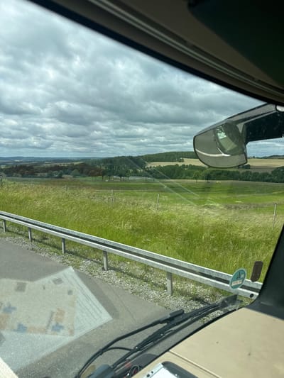 The image is taken from inside a vehicle, looking out through the windshield. The scene depicts a cloudy sky over a rural landscape with rolling hills, trees, and fields of tall grass. A metal guardrail runs along the side of the road in the foreground. The reflection of the vehicle's interior, including parts of the dashboard and windshield wipers, is visible on the road surface. In the middle distance, there are several cars on what appears to be a road or highway. There are also some distant windmills visible on the horizon. The overall impression is of a journey through the countryside on a cloudy day. The location is not specifically Plauen, Germany, but a rural area.