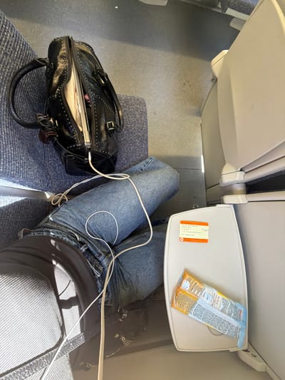 An overhead shot captures a person's lap adorned with blue denim jeans and a dark gray ribbed top, with white charging cables draped across their legs. To the left, a black studded leather bag rests on a blue upholstered seat. The bag is partially unzipped, revealing a laptop and other items within. In the foreground, a tray table attached to a beige seatback displays two packaged snacks. The floor is a textured gray, showing a faint stain. This scene is likely on a train within the Mid Sussex, United Kingdom area, given the tray table design and the presence of a train ticket visible on it.