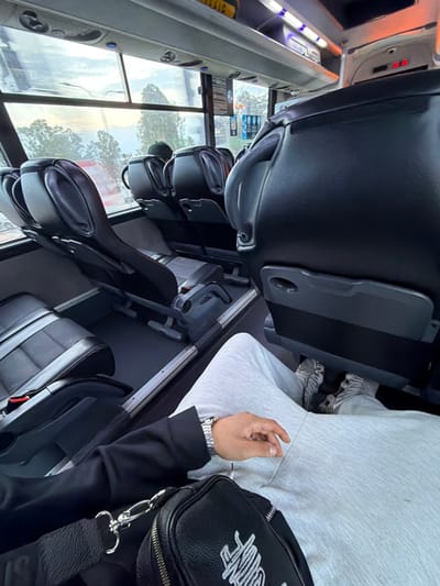 The image displays the interior of a passenger bus, observed from a first-person perspective, situated in Nesher, Israel. The immediate foreground features the legs of an individual wearing light grey sweatpants and grey sneakers. A black long-sleeved garment covers the left arm, with a silver-colored watch visible on the left wrist. A black crossbody bag, equipped with silver-toned hardware and white stylized text, rests on the lap. Ahead, a series of black leather-like passenger seats with grey fabric cushions are arranged, facing forward. The seat directly in front of the observer is unoccupied, featuring a dark grey back with an integrated fold-down tray. Further down the aisle, additional rows of seats are visible, with the head of another passenger, wearing a dark cap, discernible on the left side of the bus. Large windows on the left reveal an exterior view consisting of green trees and an overcast sky. The bus interior is illuminated by various lights, including blue overhead LEDs, and a digital display above the seats indicates "15:15" in red numerals. Metal rails run along the floor of the aisle.