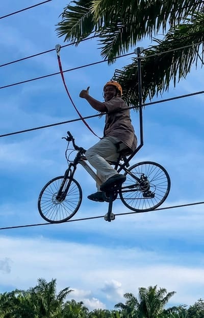 In Bintang Meriah, Indonesia, a man wearing a helmet and a brown shirt is riding a bicycle suspended from wires. The bicycle is positioned on a zipline, with its wheels resting on the cable. The man is giving a thumbs-up gesture. The background shows a blue sky with scattered clouds and palm trees. The zipline appears to be part of an adventure activity, with multiple cables running horizontally.