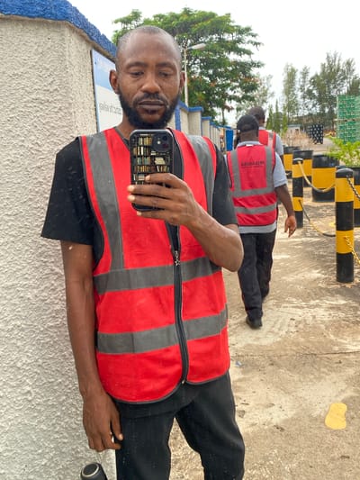 In Kinkinau, Nigeria, a man in a red and grey safety vest stands in front of a textured wall, holding a smartphone. He is wearing a black t-shirt and black pants. The phone's case displays an image of bookshelves. In the background, two other individuals wearing similar safety vests walk away from the camera. The environment appears to be an industrial or outdoor area, with black and yellow bollards lining a pathway and stacks of green-boxed items visible in the distance. The ground is a concrete or asphalt surface with some scattered debris and a yellow stain. Trees and a cloudy sky are visible in the background.