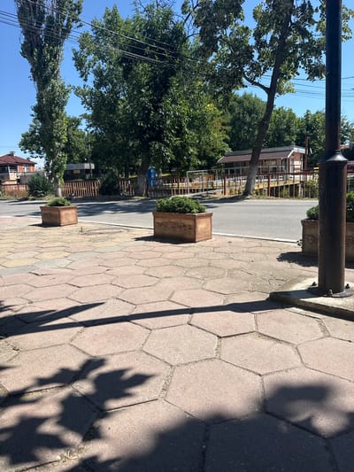 The image depicts a street scene in Shishmantsi, Bulgaria, on a clear day with strong sunlight casting shadows. In the foreground, a hexagonal-patterned sidewalk is visible, with two decorative planters containing greenery placed on it. A dark pole, likely a lamppost, stands to the right. Beyond the sidewalk, a paved road runs horizontally across the frame. On the other side of the road, a light-colored building with a brown-trimmed roof and windows is situated behind a wooden fence. Tall trees with dense green foliage line the background, and a house with a red tiled roof can be seen on the far left. The overall atmosphere is one of a quiet, residential street under bright sunlight.