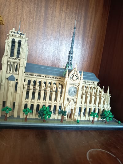 The image shows a detailed Lego model of Notre Dame Cathedral situated on a dark wooden shelf. The model features intricate architectural details, including towers, arched windows, a rose window, and a spire. The cathedral is a light tan color with a dark gray roof. Small green Lego trees with brown trunks are placed in front of the model on a gray base that simulates a sidewalk. The background is a dark wood grain texture, suggesting the model is displayed in a home or museum setting. There are no people or animals present in the image. The scene is static and depicts a constructed miniature replica. The location is described as Millstreet, Ireland, but this information cannot be visually verified from the image itself.