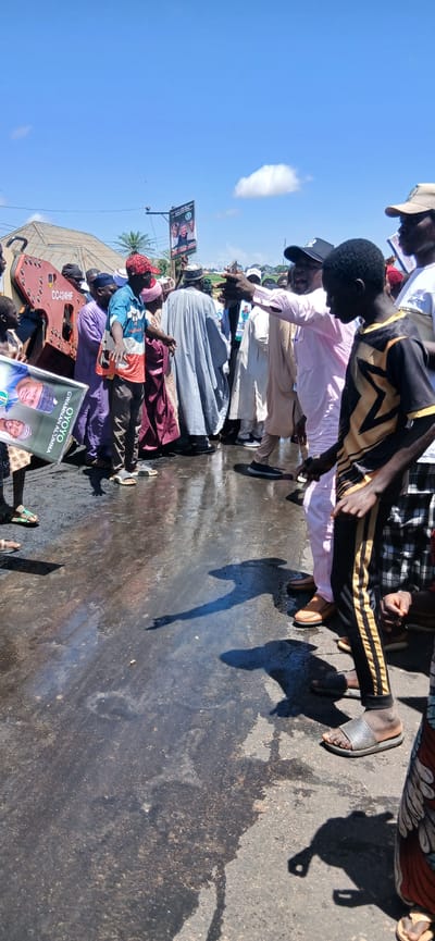 In Kayarda, Nigeria, under a clear blue sky with scattered clouds, a group of people are gathered on a wet asphalt surface. The scene appears to be outdoors, possibly during a public event or gathering. In the foreground, a young boy in a black and gold shirt and pants stands facing right, with his hands clasped. To his left, a man in a light pink garment and a cap points to the right, engaged in conversation with others.
A large poster held by individuals on the left displays images of two men and text, potentially political campaign material. Behind them, a red piece of machinery with the inscription "CC424HF" is visible. Several men are dressed in traditional attire, including caps and flowing robes. The wet ground reflects the sky and the figures, suggesting recent rain or cleaning. The overall atmosphere seems active, with people interacting and observing. A banner with images of men and text is suspended in the background, further indicating a public gathering or event.
