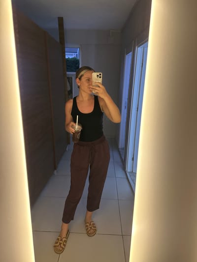 A woman is taking a selfie in a mirror. She is wearing a black tank top, maroon pants, and sandals. She is holding a drink with a straw in her left hand. The background appears to be a room with a skylight and tiled floor. Based on the location provided (Köyceğiz, Turkey), the interior setting is likely a private residence or vacation rental.