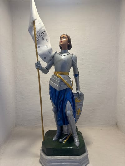 The image shows a statue of Joan of Arc, a historical figure. She is depicted as a warrior, wearing a suit of armor and holding a flag. The statue is standing on a base, which is on a shelf. The background is a white, textured wall. The statue is detailed, with realistic features. The flag has text and symbols on it. It is not possible to determine the location in Niterói, Brazil from the image.