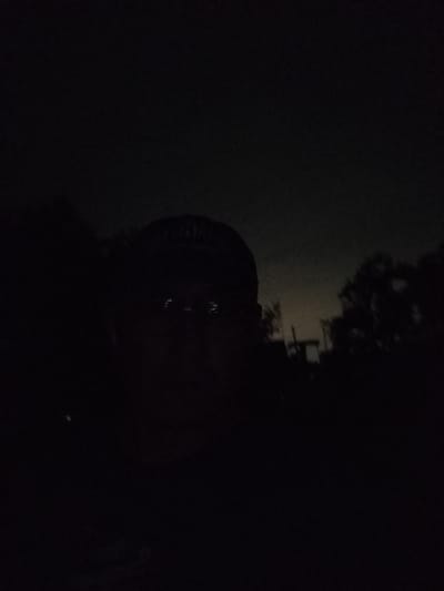 The media file is a low-light, self-portrait photograph taken outdoors. The primary subject is a person, seen from the chest up, wearing a baseball cap. Their face is dimly lit, with the eyes appearing to reflect light, suggesting the presence of a flash or a light source directed at them. The background is dark and appears to be a natural setting with trees and possibly some indistinct structures, suggesting a rural or semi-rural location. The overall atmosphere is moody and somewhat mysterious due to the low light conditions. The time of day is clearly nighttime or very early morning before dawn, given the darkness. There are no visible text elements in the image. The context provided places the location in Қарғалы, Kazakhstan.