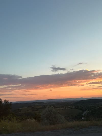 The media file depicts a landscape scene at sunset. The sky is a gradient of pale blue to a warm orange, filled with dramatic clouds catching the last rays of sunlight. The clouds are tinged with shades of pink and gold, creating a beautiful, ethereal effect.
To the right of the sky, a dark, forested hillside slopes down towards a body of water. The trees are dense and green, suggesting a natural, undeveloped area. The water's surface is rippled, reflecting the colors of the sunset. In the distance, layers of mountains fade into a hazy blue, indicating a vast and expansive landscape.
Scattered on the hillside, glimpses of human activity can be seen. There are a few buildings, possibly homes or farm structures, and some evidence of roads or paths winding through the terrain. The presence of these structures suggests a rural or semi-rural setting.
The overall mood is serene and peaceful, characteristic of a sunset. The time of day is clearly evening, as indicated by the warm, low-angled light and the vibrant sunset colors. The weather appears clear, with the clouds being the only atmospheric feature of note.
The location context provided is Golemo Buchino, Bulgaria, which helps to place this natural beauty within a specific geographical area. There is no visible text in the media file.