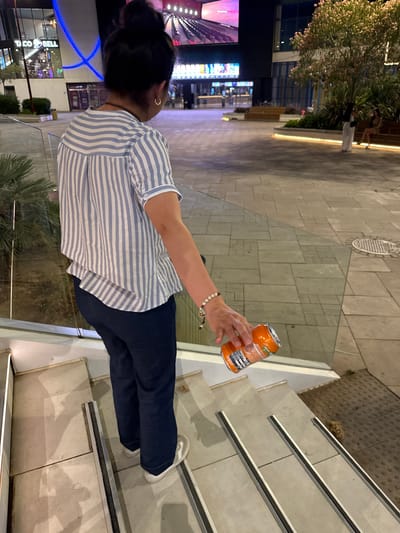 A person is standing on a set of outdoor stairs at night, holding an orange soda can. The person is wearing a blue and white striped shirt and dark pants. They have dark hair tied back and are wearing a small gold hoop earring. The background shows an outdoor area with a glass railing, tiled flooring, and some trees. In the distance, there are illuminated signs for businesses, including "TACO BELL". The scene suggests an urban or commercial setting. The lighting indicates it is nighttime.