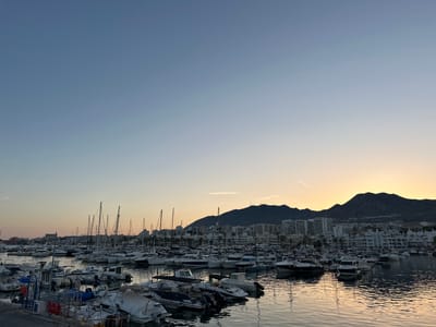 A wide shot captures a marina filled with numerous boats of varying sizes, from small motorboats to larger sailboats, docked in calm waters under a clear, gradient sky. The sky transitions from a light blue at the top to a warm orange and yellow hue near the horizon, indicating either sunrise or sunset. Silhouetted against the colorful sky are dark, rolling hills and a city skyline with numerous buildings, suggesting an urban coastal location. The water reflects the sky and the boats, creating a mirror-like effect. The foreground shows a walkway or pier with some visible boat equipment and possibly some barriers. The overall scene depicts a peaceful and picturesque view of a marina at dusk or dawn. The location is Arroyo De La Miel-benalmadena Costa, Spain. There are no visible people or discernable actions.