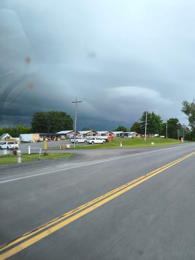 The image depicts a rural scene with a road in the foreground and a cluster of buildings in the background under a dark, stormy sky. The road is asphalt with double yellow lines. To the right of the road, a grassy embankment leads up to several buildings that appear to be a roadside market or farm stand. The buildings are rustic in appearance with wooden siding and some have awnings. Several vehicles, including white vans and trucks, are parked in front of the buildings. A utility pole with wires is visible. Trees surround the buildings. The sky is dominated by heavy, dark clouds, suggesting an impending storm. The scene is likely located in a rural area of Ontario, Canada, possibly near Alfred and Plantagenet. There are no discernible people in the image.