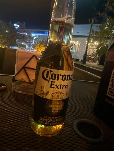A Corona Extra beer bottle lies on a metal grate surface at night. The bottle's label is prominently displayed, showcasing the brand's name and "LA CERVEZA MAS FINA" tagline. Reflections from surrounding lights glimmer on the bottle's glass. A napkin holder and a small ash tray are also visible. In the background, illuminated buildings and trees contribute to the outdoor ambiance. A Voodoo product with a QR code is placed to the right of the beer.