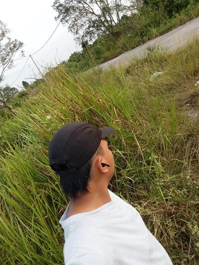 A medium shot captured from a slightly elevated angle shows a person from the shoulders up, facing away from the camera and looking upwards towards the right. The person is wearing a black baseball cap and a white t-shirt. A black wireless earbud is visible in their right ear. They are positioned amidst tall, dry grass and vegetation, with a paved road winding uphill in the background to the right. Trees with sparse foliage are present on the left side of the image, and a utility pole with wires is also visible in the upper left quadrant. The sky is overcast and bright. The overall scene suggests an outdoor environment, possibly a roadside or hillside. The presence of the road and vegetation indicates a rural or semi-rural setting, potentially in or near Kualu, Indonesia.