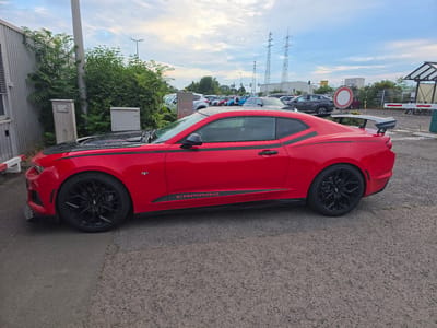 A red Chevrolet Camaro ZL1 is parked on an asphalt surface in a lot. The car features black accents on the hood and side skirts, along with black wheels. In the background, there are other vehicles, electrical towers, and some vegetation. The sky is overcast. The location is identified as the Worms, Germany area.