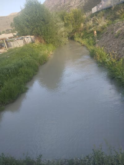 An elevated shot shows a wide, muddy river flowing through a rural or semi-rural landscape. The river occupies the majority of the frame, with a slight ripple on its surface. The banks of the river are lined with lush green grass on the left and a dry, dusty hillside with sparse vegetation on the right. In the background, a range of barren mountains is visible under a clear sky.

On the grassy bank to the left, there are some low-lying structures, possibly sheds or agricultural buildings, partially obscured by trees. On the right bank, further up the hill, a modern metal pedestrian bridge spans across an elevated path. Several people are visible on the bridge, appearing to be walking or standing. Closer to the river on the right bank, a single, light-brown animal, possibly a dog or a small deer, is seen standing on the grassy slope.