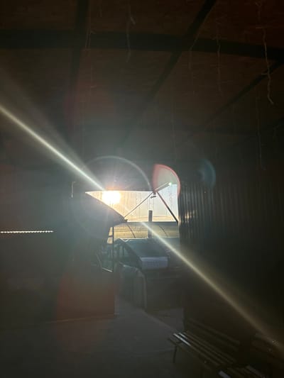 This image captures a dramatic interplay of light and shadow within a dimly lit interior space. The primary subject is the bright sun, intensely shining through an opening, creating strong lens flares and a powerful beam of light that diagonally cuts across the frame. The opening appears to be a window or doorway, offering a glimpse of the bright sky outside, possibly suggesting a time of day around sunrise or sunset given the warm, golden hue.
The interior setting is rustic and somewhat disheveled. Walls appear to be constructed from rough wooden panels, and there are visible metal beams and supports. Delicate strings of what look like fairy lights are strung across the upper parts of the interior, adding a touch of decoration but also contributing to the cluttered feel. In the foreground and midground, there are hints of objects, including what might be a mattress or bedding on the left and some stacked items or furniture on the right. The overall impression of the interior is one of an unfinished or storage space, possibly a workshop, shed, or even a temporary shelter.
The intense sunlight suggests fair weather outside, and the scene evokes a sense of drama and perhaps a touch of mystery due to the strong contrast between the illuminated opening and the shadowed interior. The location is identified as Заплавская, Russia. There is no visible text in the image.