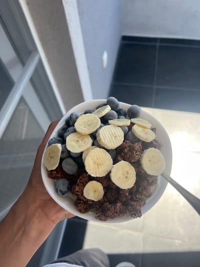 In a white bowl, a serving of cereal is presented with sliced bananas and blueberries. The cereal appears to be a granola or muesli mix, dark brown in color, with some pieces slightly larger and more clustered. Scattered throughout are vibrant blue blueberries, some appearing slightly frosted. Several slices of banana, showing their characteristic circular shape and subtle ridges, are arranged on top of the cereal and fruit. A silver spoon is visible, partially submerged in the bowl, suggesting the cereal is about to be eaten.

The bowl is held by a person's hand, with a hint of their arm and shoulder visible on the right side, dressed in a light blue shirt. The background suggests an indoor setting with strong natural light casting shadows, possibly indicating a bright, sunny day. Visible in the background are dark tiled walls and a light-colored countertop or table surface, with some reflections suggesting glass or a shiny surface. The overall impression is of a healthy and refreshing breakfast or snack, enjoyed in a well-lit environment. The location context of Chaidefto, Greece, suggests a potentially warm climate, and the bright lighting aligns with this. No visible text is present in the image.