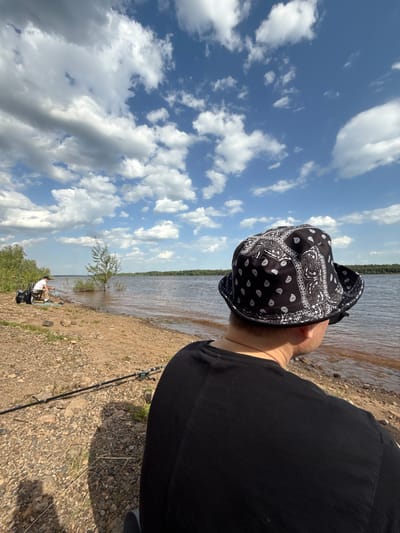 A person wearing a black bandana bucket hat and a black t-shirt is standing on a rocky shoreline looking at a body of water under a sky filled with clouds. In the background, another person is sitting on a chair with fishing equipment nearby. The photo was taken near Doksha, Russia.