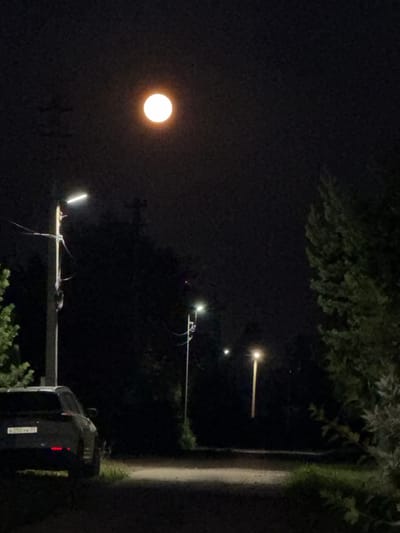 A nighttime scene in Simonikha, Russia, is captured with a bright moon dominating the dark sky. In the foreground, a dark-colored SUV is parked on the right side of a dirt road. The car's license plate, partially visible, reads "B255YK 37". To the left of the road, a street light illuminates a portion of the area. Further down the road, another street light is visible, casting a warm glow. The surroundings are filled with dense foliage and trees, indicative of a rural or semi-rural setting. The overall atmosphere is serene and quiet, with the moonlight and streetlights providing the only sources of illumination.