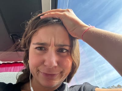 A medium close-up, slightly low-angle photograph depicts a young female subject inside what appears to be a vehicle. The subject has medium-brown hair, dark eyes, and a facial expression characterized by furrowed eyebrows and slightly downturned lips, suggesting bewilderment or mild discomfort. Her right hand (viewer's left) is raised, with fingers resting on her forehead and hair. A pink string bracelet is visible on her right wrist. She is wearing a dark, possibly ribbed, top, and white wired earbuds are inserted into both ears, with wires visible. A small gold hoop earring is present in her left ear. The vehicle's interior is partially visible on the left side of the frame, showing a dark overhead structure and glimpses of red-patterned and light-textured fabric, possibly upholstery or curtains. The right side of the image reveals a bright blue sky with scattered wispy white clouds, viewed through a window. The specific location within Leiria, Portugal, cannot be determined from the visual evidence provided in the image.