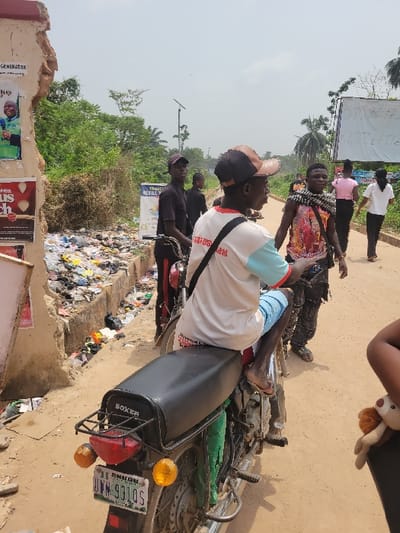 The image depicts an outdoor street scene located in Enugu, Nigeria, as indicated by the 'ENUGU' license plate on the motorcycle, contrary to the prompt's reference to Candelaria, Colombia. The unpaved dirt road, under a hazy sky, shows visible individuals and objects.

In the immediate foreground on the right, a partial view of a dark-skinned individual's arm and shoulder is visible, holding a small, light brown stuffed animal. Central to the scene, a dark-skinned male, wearing a white t-shirt with light blue sleeves and red trim, light blue shorts, and brown sandals, is seated on a black 'BOXER' brand motorcycle. He wears a brown and black baseball cap backward and has a black strap across his left shoulder. The motorcycle features a black seat, a green rear mudguard, red taillight, two amber turn signals, and a black rear rack displaying the license plate 'ENUGU UWH-931QS'. The seated man faces right, away from the camera.

To the left of the motorcycle, a dark-skinned male in dark clothing and a cap stands near a bicycle or another motorcycle, also facing right. Behind him, another dark-skinned male, largely obscured, is visible. To the right of the seated motorcyclist, a dark-skinned male, wearing a colorful sleeveless shirt with patterns and dark trousers (possibly cargo pants with chains), walks forward while looking towards the camera with a slight smile. In the background on the right, two dark-skinned females walk away from the camera: one in a light pink top and dark pants, and the other in a white top and dark pants.

The environment on the left features a dilapidated, light-colored wall, revealing exposed reddish-brown structural material. Several posters are affixed to the wall, including one with a portrait of a smiling man in a green shirt and another red poster with white text. Adjacent to the wall, a ditch or gutter is filled with a substantial accumulation of trash, including plastic bags, bottles, and general refuse. On the right and in the background, dense green vegetation, including palm trees, lines the road. A white sign with blue text "TRANC REFILLS" is visible in the mid-ground, and further right, a large white sign or billboard displays dark, handwritten numbers. A solar-powered street light on a pole stands in the background.