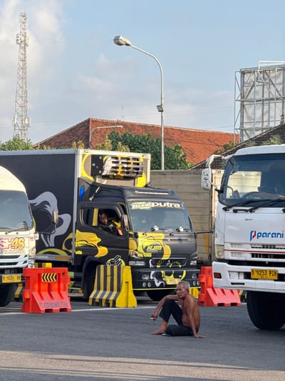 The image depicts a street scene in Melaya, Indonesia, where several large trucks are parked. In the foreground, a man is sitting on the ground, shirtless, wearing dark pants, and holding a cigarette. He appears to be looking towards the left side of the frame. To his right, a white truck with "parani" logo is visible. To his left, a black and yellow decorated truck with a driver inside the cabin is present. Traffic barriers, one orange and one black and yellow striped, are placed on the road. In the background, a cell tower and buildings with tiled roofs can be seen under a clear blue sky. The overall atmosphere suggests a roadside setting, possibly a truck stop or a waiting area.