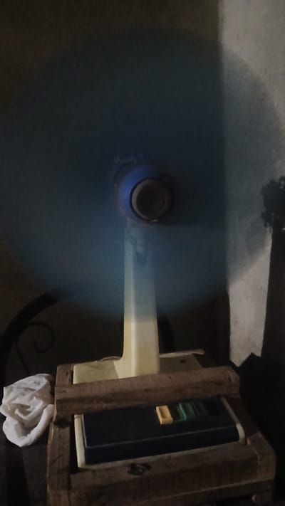 The image shows a spinning circular object, likely a fan or rotating machinery, positioned on a dark surface. A wooden frame supports a blue electronic device, possibly a control panel or monitor, with visible illuminated segments. A white cloth is seen partially obscuring the bottom right corner of the frame. The location in Putucual, Venezuela is not apparent from the image.