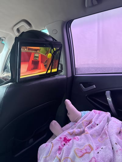 This media file captures an interior view of a car, looking towards the rear seat and out a sunroof or large window on the left. The scene appears to be set during the day, with bright, diffused light visible through the windows, casting a distinct purple hue, strongly suggesting the car is inside an automated car wash tunnel.
In the back seat, a child or infant is partially visible, covered by a light pink blanket with indistinct patterns. Only their feet, clad in matching light pink socks, are clearly seen. Mounted to the back of the front passenger seat headrest is a tablet, secured with black elastic straps. The tablet screen displays a vibrant, colorful cartoon or animated scene, featuring red structures, yellow circular objects, and other bright elements. The back of a person's head, likely an adult, is partially visible in the lower center-right, showing dark hair with some lighter strands and a patterned head covering or headband.
The car's interior features black leather seats and dark plastic trim, contrasted with lighter gray or beige headliner and grab handles. A car door with a silver handle accent is visible on the upper right. The overall impression is one of a comfortable journey, with the child entertained by the tablet during a car wash. The specific location, Kiselevka, Russia, is provided, though no visual cues within the image confirm this detail. There is no discernible text visible in the image.