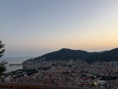 An elevated panoramic view captures the coastal city of Budva, Montenegro, nestled against a backdrop of lush, dark green mountains. The sky transitions from a pale blue at the top to a soft, warm gradient of yellow and orange near the horizon, indicating either sunrise or sunset. A faint crescent moon is visible in the upper right portion of the sky, accompanied by a small, bright star.

The city sprawls across the hills, a dense collection of buildings with red-tiled roofs interspersed with taller, modern structures. Lights are beginning to illuminate the city, casting a glow on the urban landscape. Along the coast, a bay is filled with numerous boats and yachts, their lights reflecting on the calm water. The shoreline is a mix of urban development and natural terrain.

In the foreground, a dark metal railing runs horizontally across the bottom of the frame, suggesting the viewpoint is from a road or a raised platform. To the left, the dark silhouette of pine trees partially obscures the view of the city, framing the scene. The overall impression is one of a tranquil evening over a bustling coastal city.