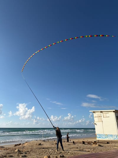 The scene depicts a beachfront environment at Beit Yanai, Israel, under a clear blue sky with scattered white cumulus clouds.
In the foreground, a person with long hair, dressed in a dark jacket and blue jeans, stands on a sandy beach. This individual is holding a long, dark, pole-like object that extends diagonally upwards and left, tethering a very long, multi-segmented kite. The kite displays a rainbow color pattern (red, orange, yellow, green, blue, purple) and arcs across the upper portion of the sky. To the right of the primary individual, a second person in dark clothing stands further back on the sand, appearing to look down at an object in their hands.
The beach itself consists of sand interspersed with dark rocks and debris, transitioning to the Mediterranean Sea with moderate waves breaking. One individual on a board is visible in the water in the mid-ground. Extending into the sea in the background is a dark, linear structure, possibly a pier or breakwater.
On the right side of the image, a two-story, white wooden structure resembling a beach hut or lifeguard station stands on the sand. It features boarded-up windows on the upper level and a blue and white wave-like design painted along its lower facade. A reddish-brown wooden planked surface is partially visible at the bottom right corner.