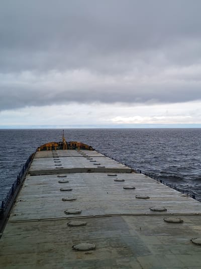 The image shows the deck of a ship at sea under a cloudy sky. The deck is flat and gray, marked with evenly spaced circular hatches. The ship is heading towards the horizon, where a faint band of light separates the water from the sky. The water is dark and choppy with small waves. There is no indication of the city of Prislon', Russia in the image.