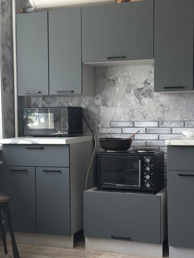 The image depicts a modern, minimalist kitchen with grey cabinetry and a grey and white marbled backsplash. On the countertop, to the left, sits a black microwave oven with a reflective glass door displaying a reflection of trees and sky, with an Apple logo visible on the screen. Beneath the microwave is a grey cabinet with two drawers. To the right of the microwave, there is a smaller black oven on top of a grey cabinet, with a black frying pan and a wooden spoon placed on top of the oven. The overall environment is clean and organized. There are no people visible in the image. The location is specified as Yapykovo, Russia.