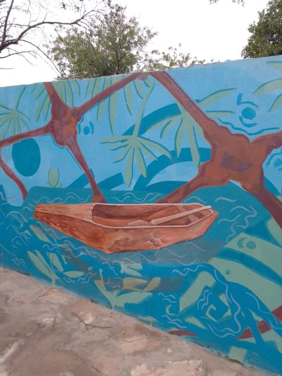 The image depicts a mural on a wall in Villa Hayes, Paraguay. The mural features a brown dugout canoe on a light-blue background representing water. Stylized waves are depicted in shades of blue and green. Above the canoe, stylized trees with brown trunks and green palm-like foliage are painted. In the background, the sky is visible with some trees. A concrete base of the wall is seen along the bottom edge of the image.