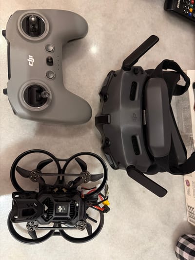 The image displays a DJI drone, goggles, and controller arranged on a light-colored, textured surface. The drone is positioned at the bottom, its black frame and propellers clearly visible. Above it, the dark grey goggles with a black strap are situated. To the left of the goggles and above the drone, a light grey controller with joystick controls and buttons is placed. A black remote control is partially visible in the upper right corner of the image, along with a portion of a white box with text and a black and white checkered fabric item. The scene is lit, showing distinct shadows cast by the objects. No people are present in the image, and there is no indication of the city of Ban Phonsavang Neua, Laos.