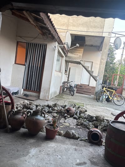 The image displays an outdoor residential courtyard or alleyway in Novi Pazar, Bulgaria, characterized by concrete buildings and rustic elements. The perspective looks down into the space.
On the left, multi-story buildings constructed from concrete rise, with some sections exhibiting exposed rebar, suggesting an informal or perhaps unfinished style. Various windows are visible, some fitted with dark, striped blinds, while others show an orange hue from internal light or reflection. Several white satellite dishes are mounted on the rooftops and walls, interconnected by visible cables. A section of a brown-tiled roof is also apparent on one of the buildings.
The ground on the right features a concrete pathway, interspersed with soil and a border of irregular, light-colored rocks. Along this rocky border are several large, aged, terracotta-colored ceramic pots or amphorae, one of which contains a small potted plant. In the immediate foreground, parts of a wooden barrel with distinctive red bands and a red wooden wagon wheel are visible.
Further into the scene, a pair of dark flip-flops rests on a wooden plank near a doorway. Two bicycles are present: a dark-colored bicycle with "BOGATTI" visible on its frame leans against a low wooden bench or step, and a yellow mountain bike, with "SPRINT" visible on its frame, rests against a metal gate at the top of a concrete outdoor staircase with wooden treads.
The scene is lit by natural daylight, indicating it is daytime. No specific human activity is depicted; the area appears lived-in and functional. The architectural style, the array of satellite dishes, and the traditional decorative items like the pots and barrel offer a glimpse into the local environment.
