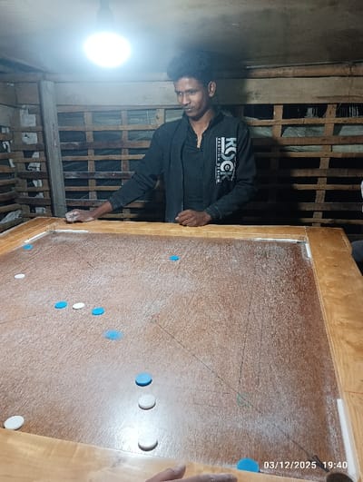 This image, captured at 19:40 on March 12, 2025, in Maliat, Bangladesh, depicts an indoor scene featuring a carrom game. A young adult male, with dark, curly hair and a medium complexion, is positioned behind a large, light brown wooden carrom board. He wears a black long-sleeved shirt or jacket with white text reading "ROCK FASHION DESIGN" on the right sleeve and "KSQ" on the chest, over a dark undershirt. His left hand rests on the far-left edge of the board, and his right hand is near the center-right edge. He looks towards the right side of the frame.
The carrom board's playing surface is light brown, marked with game lines, and dusted with a fine white powder, likely carrom powder. Numerous circular carrom coins, both white and light blue, are scattered across the board. A second hand, presumably belonging to another player, is visible in the bottom right corner of the frame, also resting on the board.
The environment appears to be a modest, enclosed space. A single bright, bare light bulb hangs from the ceiling, illuminating the scene. The background consists of rough wooden slat or pallet construction, possibly with a tarp or plastic sheeting visible behind it, suggesting a makeshift or utilitarian structure.