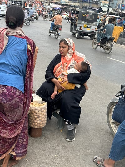 The image depicts a busy street scene in Karachi, Pakistan, likely during daytime with overcast or diffused lighting. The foreground features two women, one holding a baby. The woman in the center is wearing a black abaya and an orange and white patterned headscarf. She is holding a sleeping baby, who is dressed in a yellow garment. Beside her is a woven basket filled with what appear to be snacks, possibly nuts or fried dough. In the foreground, another woman, seen from behind, is wearing a maroon and purple patterned sari with a blue garment underneath.
The background is filled with various vehicles, including auto-rickshaws (one with "SAZGAR" text and a license plate "AAO 9679"), motorcycles, and a white car. People are seen on and around these vehicles. The setting is a paved street, indicating an urban environment, likely a marketplace or a busy thoroughfare given the density of vehicles and pedestrian activity. The overall atmosphere suggests a typical day in a bustling Pakistani city, with a focus on the everyday lives of its inhabitants. There are no explicit indicators of specific emotions, but the scene conveys a sense of daily life and commerce.