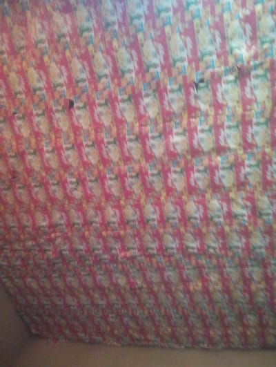 This image, located in an interior space within the city of Idon, Nigeria, presents a close-up, upward-angled view of a ceiling or upper wall covered by an improvised, patterned material. The material consists of numerous vertically oriented rectangular segments, predominantly red, arranged in a repetitive grid. Each segment features a light-colored, possibly anthropomorphic, cartoon character wearing a red hat, with yellow and blue elements in the background, resembling repurposed product packaging or printed paper. Two distinct, irregular dark holes or tears are visible in the material in the upper-central portion of the frame. The lower edge of the image reveals a plain, light brown/beige surface, likely a wall or structural element, below the patterned covering. No individuals, explicit actions, or interactions are discernible.