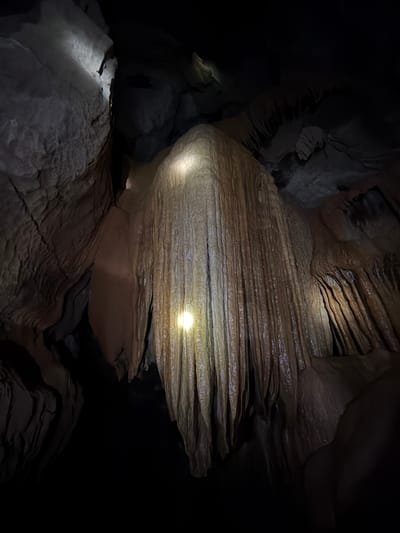 The image displays the interior of a cave in Phang-nga, Thailand, illuminated by artificial light sources. A large, multi-textured flowstone formation dominates the central frame, extending vertically downwards from the ceiling. This formation exhibits a predominantly reddish-brown to light brown coloration with visible striations and a wet, reflective surface. A bright, circular beam of light originates from the lower-center-left, directly illuminating the central flowstone. A second, more diffuse light source from the upper-left casts light upon a rugged, grey rock wall to the left of the central formation. The surrounding cave walls and ceiling are composed of dark grey to black rock, with irregular textures and shadowed crevices. No individuals or anthropogenic objects are visible within the scene.
