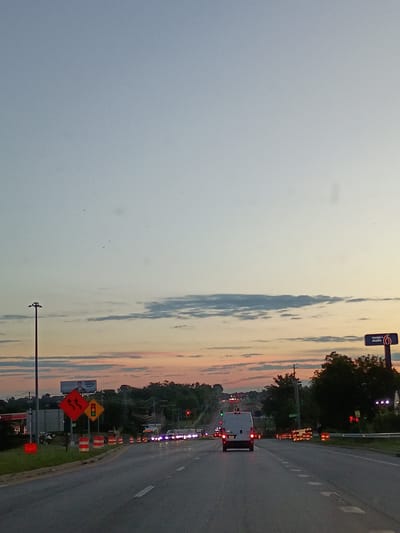 The media file depicts a street scene at dusk in Lexington, United States of America. The sky is a soft blend of blue and pastel hues, suggesting the sun is setting or has recently set. Wispy clouds are visible against the fading light.

The primary subject is a road with multiple lanes, bordered by a grassy shoulder. Several vehicles are present on the road, including a white van with its brake lights illuminated, and other cars further down the road, some with their headlights on. Traffic signals are visible and appear to be red, indicating a stop.

The scene shows ongoing road construction. Orange and white traffic cones are lined up along the shoulder and in the road, channeling traffic. Several construction signs are also present. One sign, diamond-shaped and yellow, displays traffic signals. Another, red and diamond-shaped, depicts a series of wavy lines, possibly indicating a winding road or road hazard.

A prominent object on the left side of the road is a tall streetlamp with lights. In the background, there is a billboard with an image of a person and some text, partially obscured. A sign for "Motel 6" is also visible, indicating lodging nearby. Trees and distant buildings form the backdrop.

The overall atmosphere is calm, with the gentle light of dusk and the presence of construction suggesting a transition period. The time of day is clearly evening, and the weather appears clear.