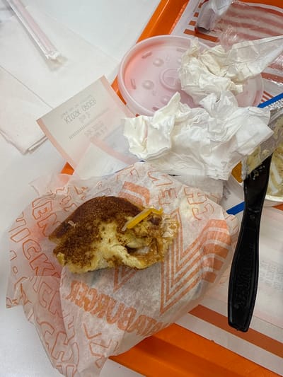 The image shows a partially eaten cinnamon roll on a Whataburger wrapper. A black plastic knife rests nearby, along with crumpled napkins, a plastic cup lid, a straw wrapper, and a crumpled kiosk order receipt. The scene suggests a fast-food meal has been consumed, leaving behind a cluttered collection of trash on an orange tray.