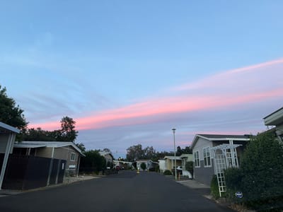 The image shows a street in a mobile home park at dusk in Citrus Heights, California. The sky is a gradient of blue and pink, with wispy clouds. The street is paved and curves to the right. On either side of the street are mobile homes of various sizes and colors, many with small yards. There are trees and some landscaping visible. A street lamp is visible in the middle of the street. The overall impression is one of a quiet residential area during the twilight hours.