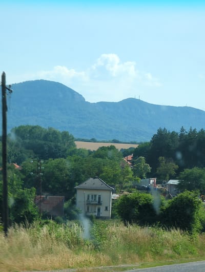 This image depicts a rural landscape under a bright, clear sky, suggesting daytime. The scene is dominated by a large, lush green mountain range that stretches into the distance. In the foreground and mid-ground, there are signs of human habitation and activity. Several houses with tiled roofs are visible, nestled amongst dense trees. A field of dry, golden crops is situated between the mountain and the cluster of houses.

On the right side of the image, a roadside scene is captured. Tall, dry grass lines the edge of a paved road, indicating a natural, possibly uncultivated area. There are hints of construction or industrial activity with some machinery and structures partially obscured by trees and vegetation. A plume of white smoke or steam is rising from an area with these structures, suggesting ongoing work or an industrial process. Power lines and poles are also visible, crisscrossing the scene and suggesting a developed but rural area. The overall impression is one of a working rural landscape, possibly on the outskirts of a village, with the natural beauty of the mountains as a backdrop. No people are clearly visible in the image, and the weather appears to be fair and sunny. There is no visible text in the image.